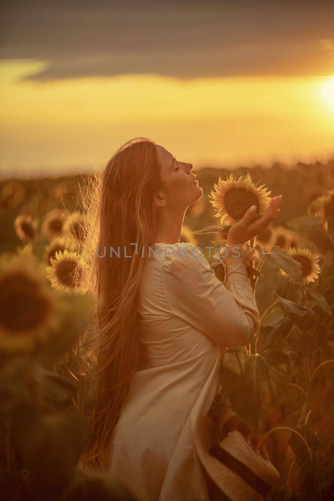 Beautiful middle aged woman looks good in a hat enjoying nature in a field of sunflowers at sunset. Summer. Attractive brunette with long healthy hair