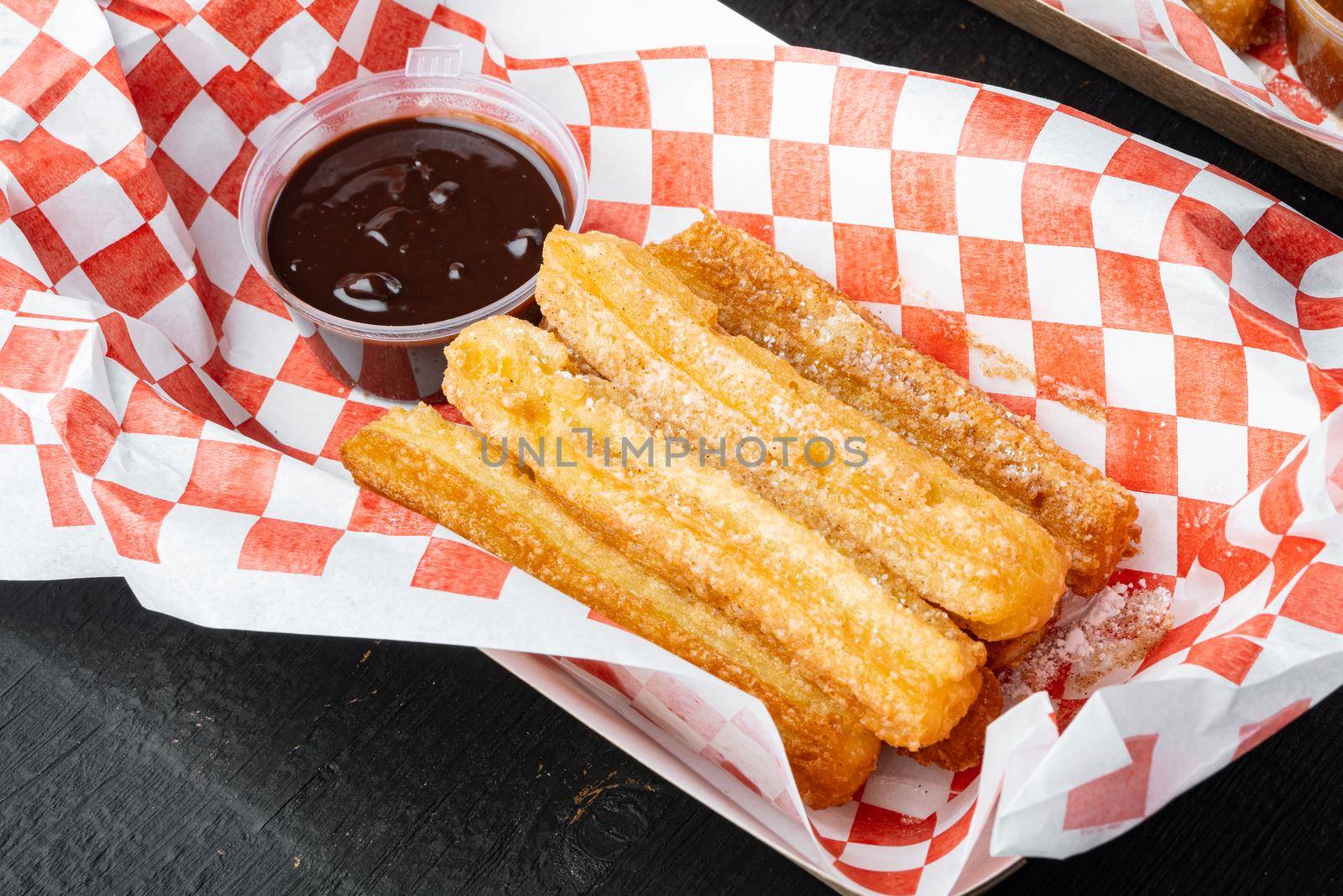 Tasty hot sweet churros in paper with chocolate, and caramel in paper tray, on black wooden table background