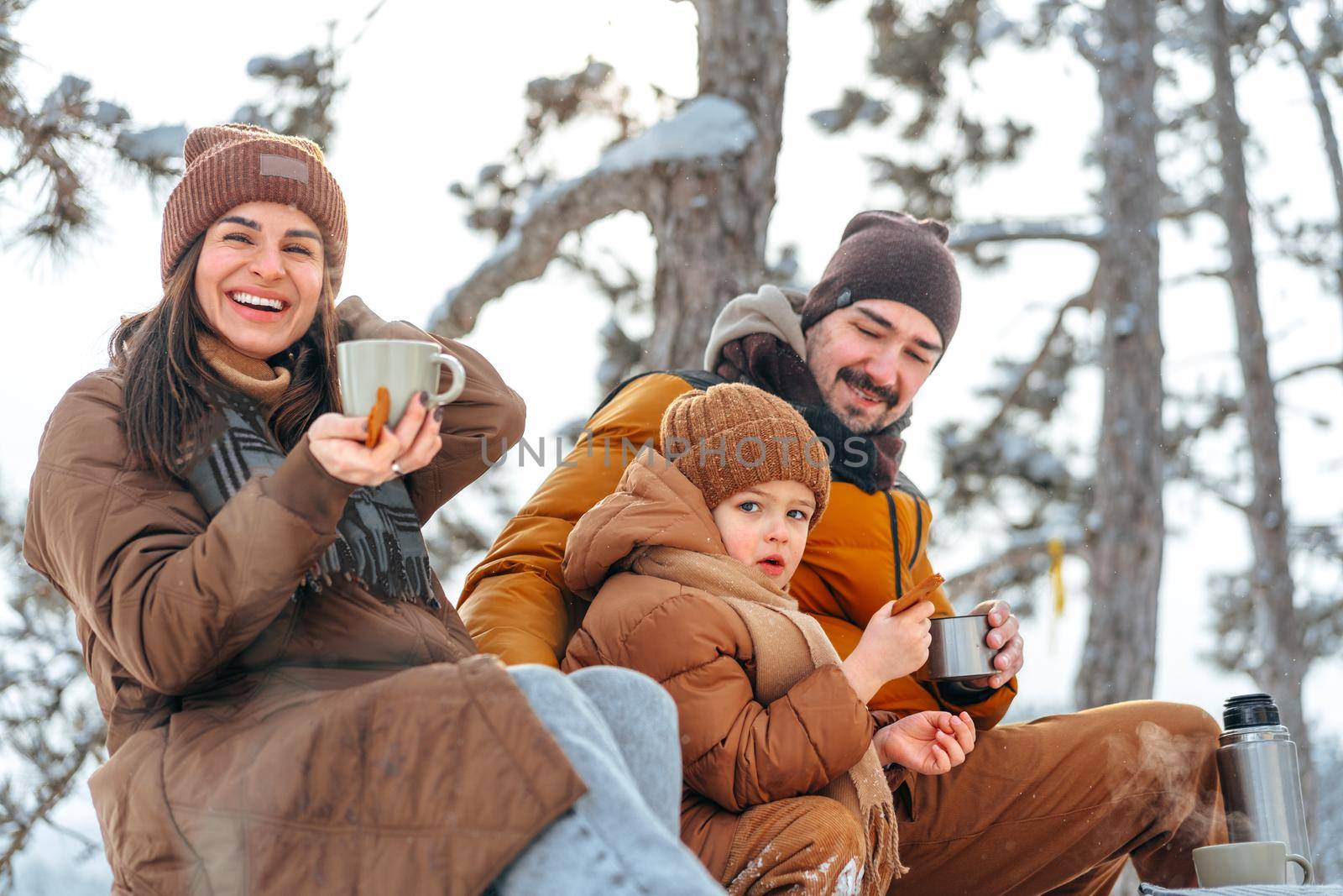 Happy family with cups of hot tea spending time together in winter forest by Fabrikasimf