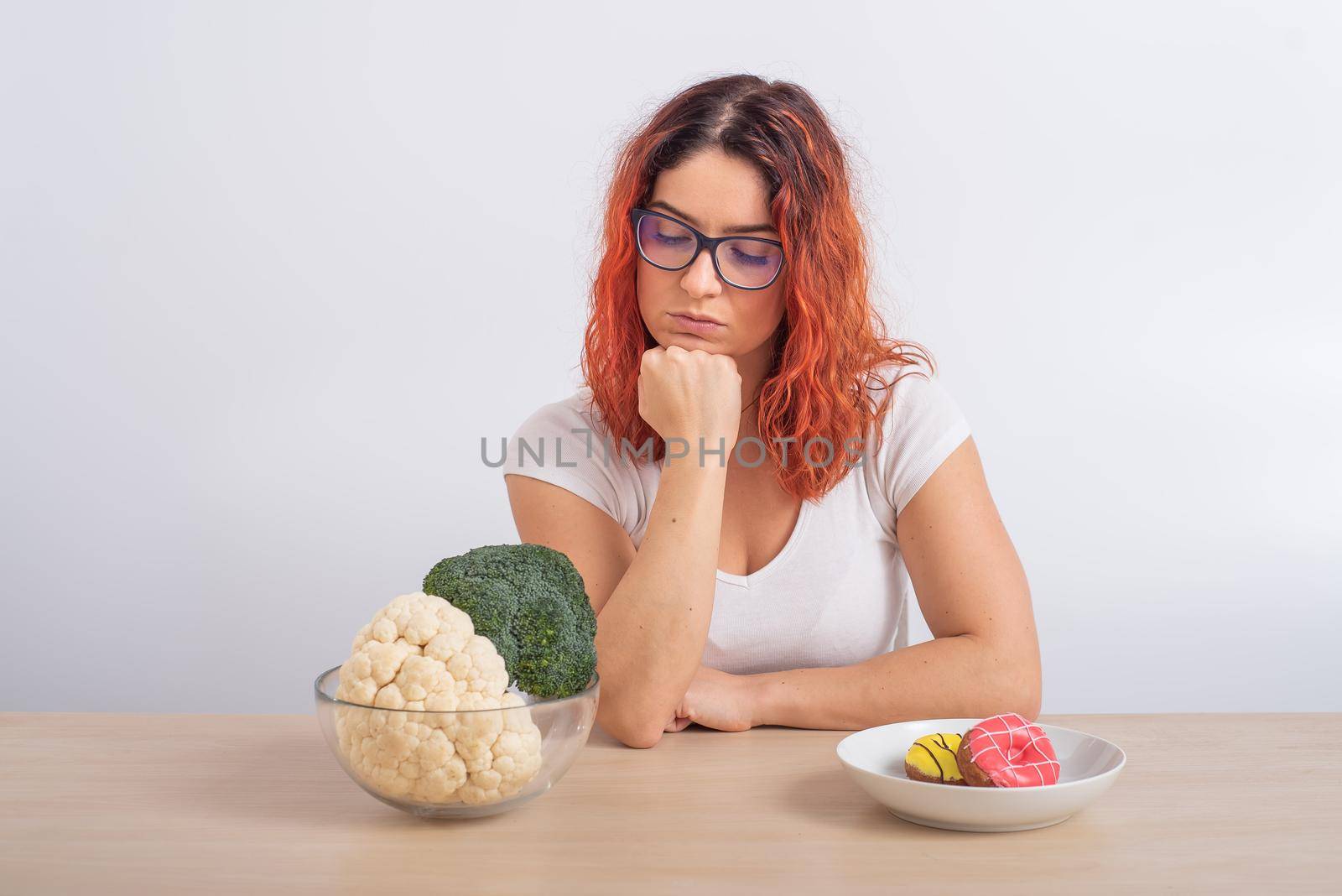 Caucasian woman prefers healthy food. Redhead girl chooses between broccoli and donuts on white background. by mrwed54