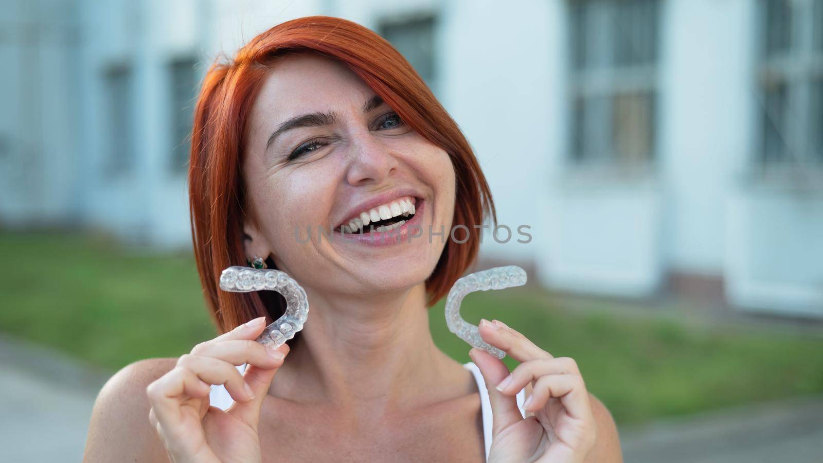 Red-haired Caucasian woman holding transparent mouthguards for bite correction outdoors. A girl with a beautiful snow-white smile uses silicone braces by mrwed54