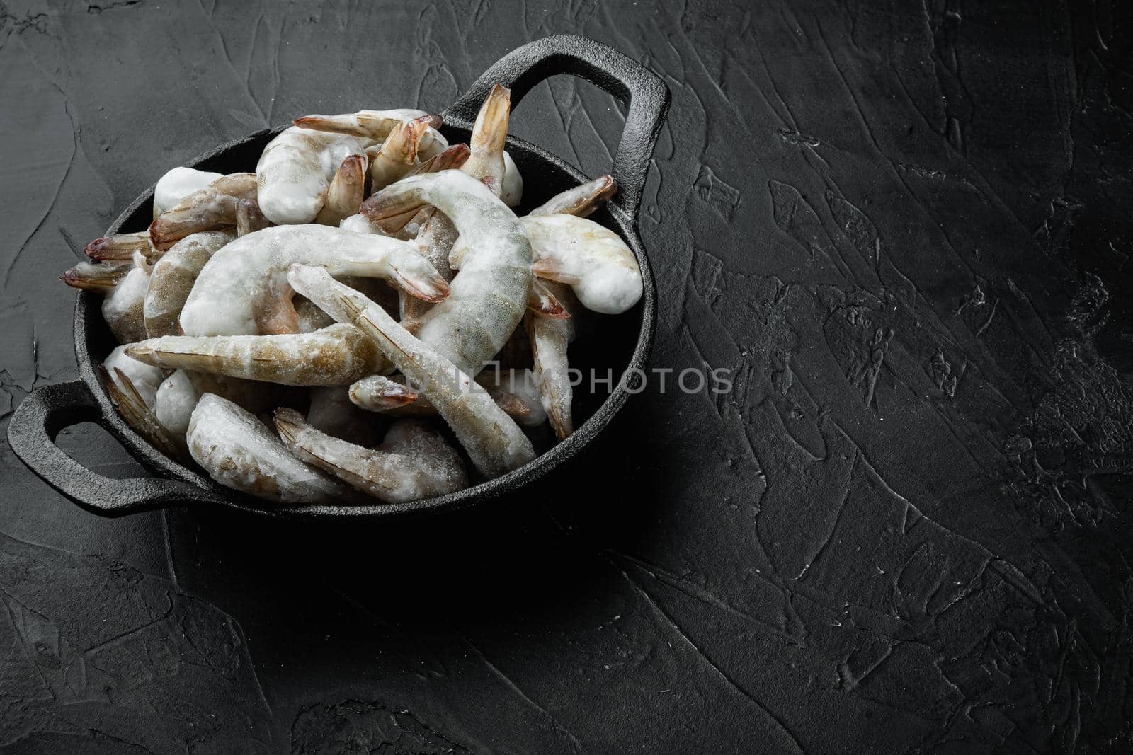 Frozen raw uncooked tiger prawns, shrimps, in cast iron frying pan, on black stone background, with copy space for text by Ilianesolenyi