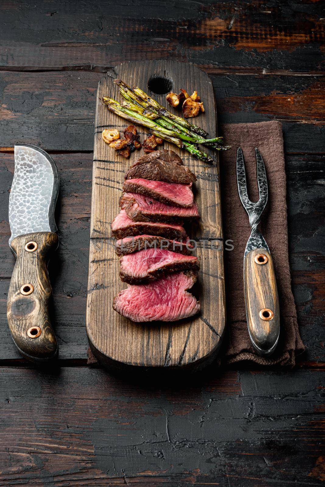 Fillet Mignon tenderloin Grilled and sliced meat beef steaks medium rare, with onion and asparagus, on wooden serving board, with meat knife and fork, on old dark wooden table background by Ilianesolenyi