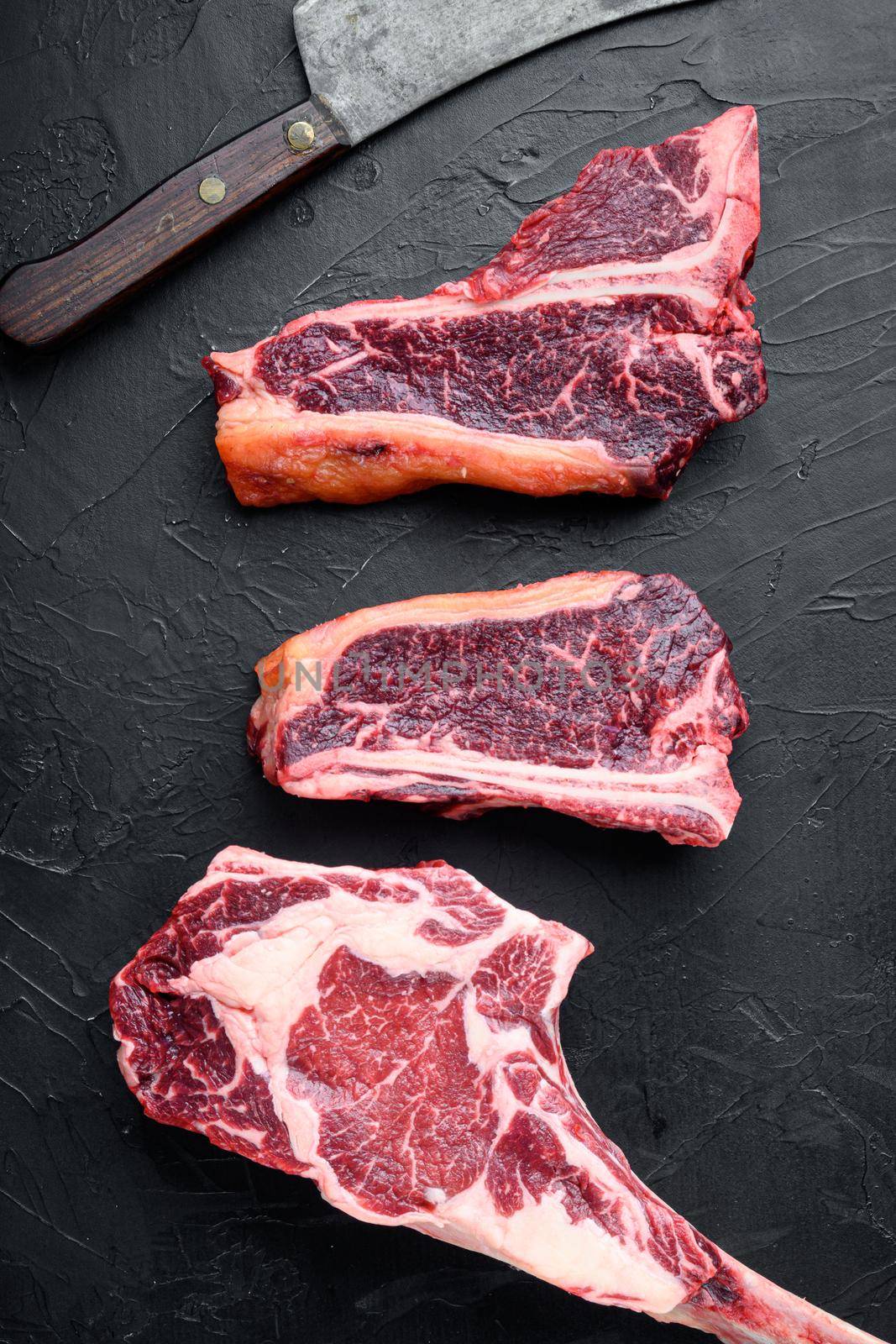 Dry aged marbled beef meat cut, tomahawk, t bone or porterhouse and club steak , with old butcher cleaver knife, on black stone background, top view flat lay by Ilianesolenyi