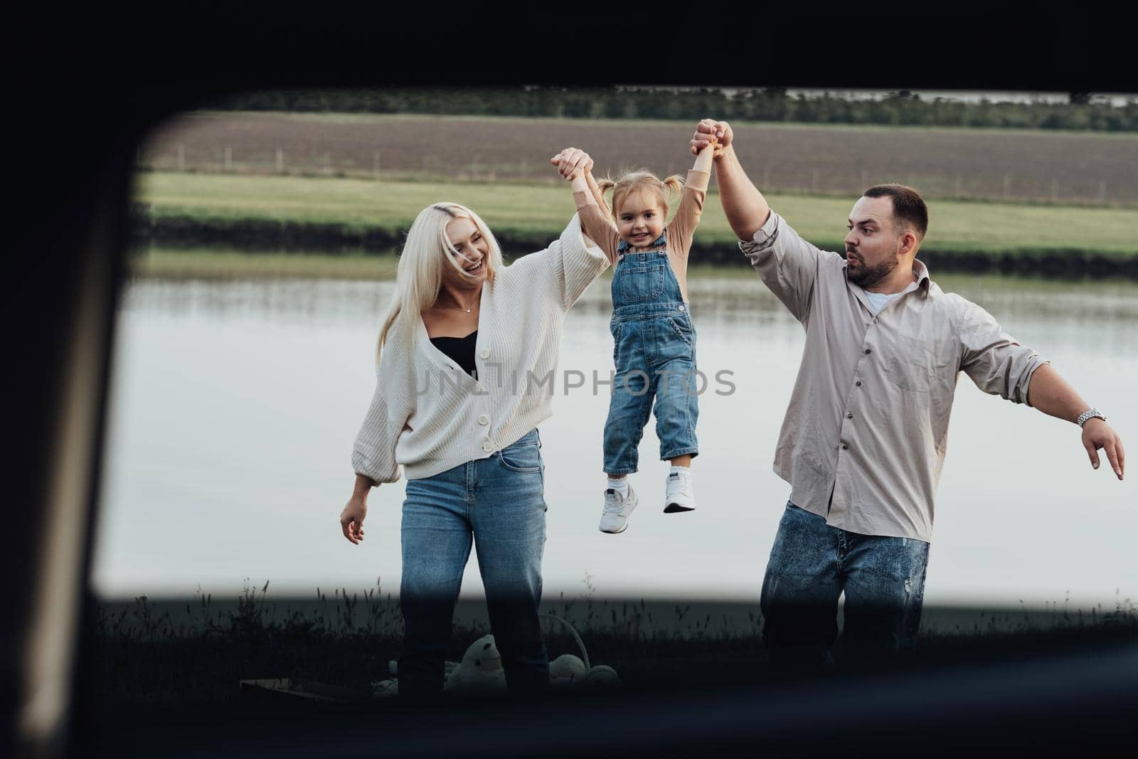 Happy Young Family Having Fun Outdoors, Mom and Dad Taking Up Their Little Daughter by Hands, View Through Window Inside of Car