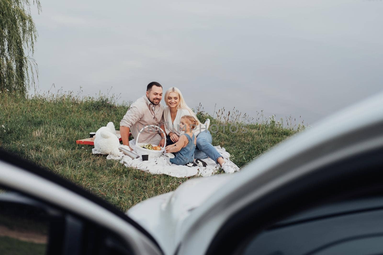 Young Family with Little Kid Enjoying Picnic in Front of Their Car, Weekend Outdoors Near Lake