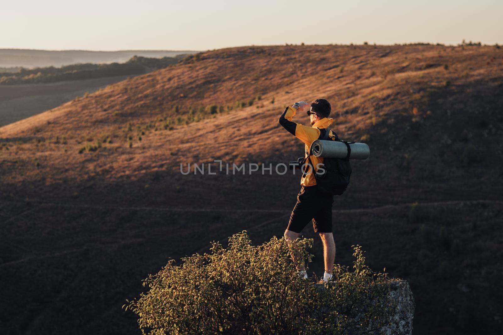 Young Man Traveler with Camera Standing on the Top of Rock and Looking Into Sunset, Male Tourist with Backpack and Mat Enjoying His Solo Trip by Romvy