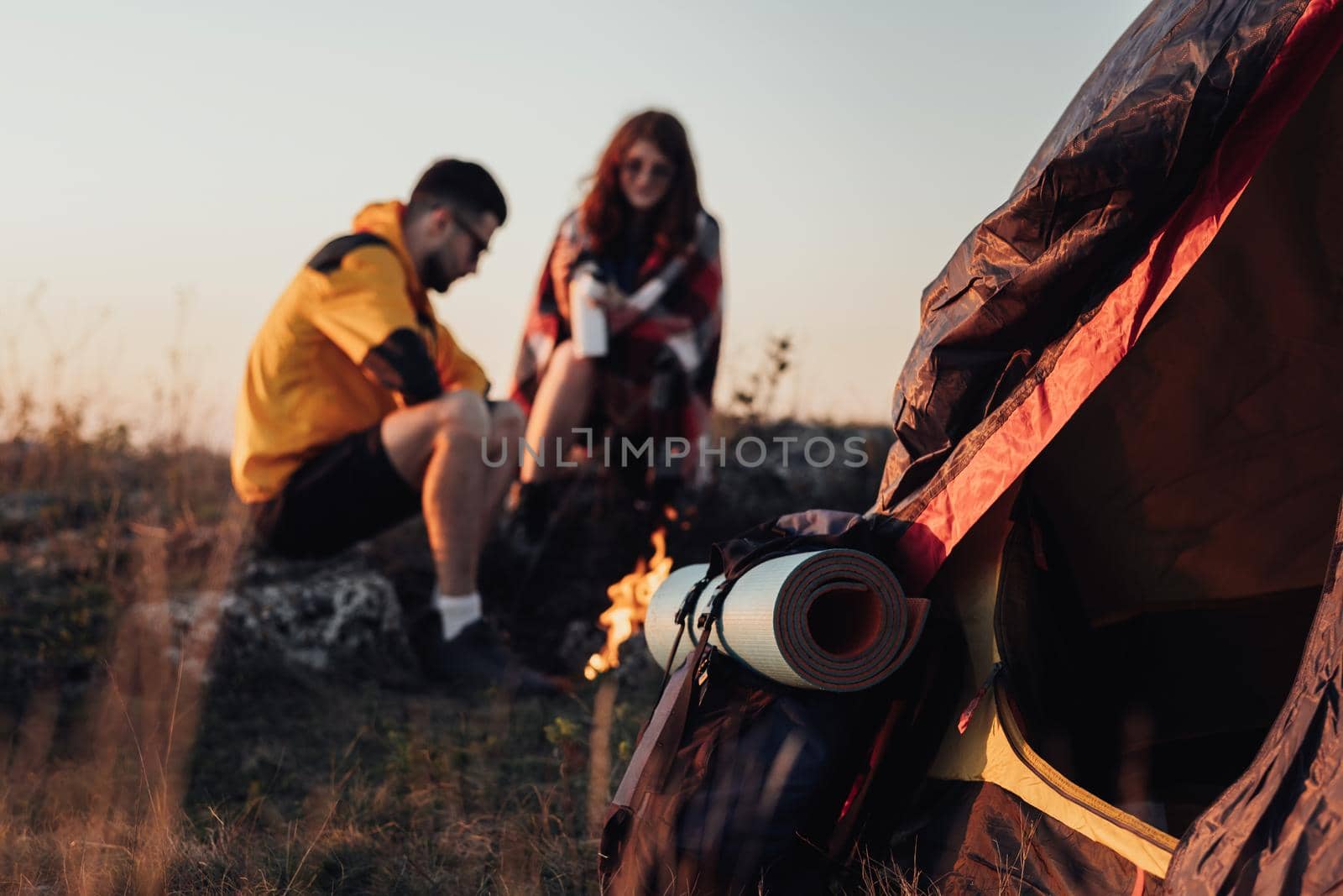 Backpack with Mat and Tent in Focus, on the Background Two Young Travelers Man and Woman Sitting Near Campfire During Sunset, Travel Concept