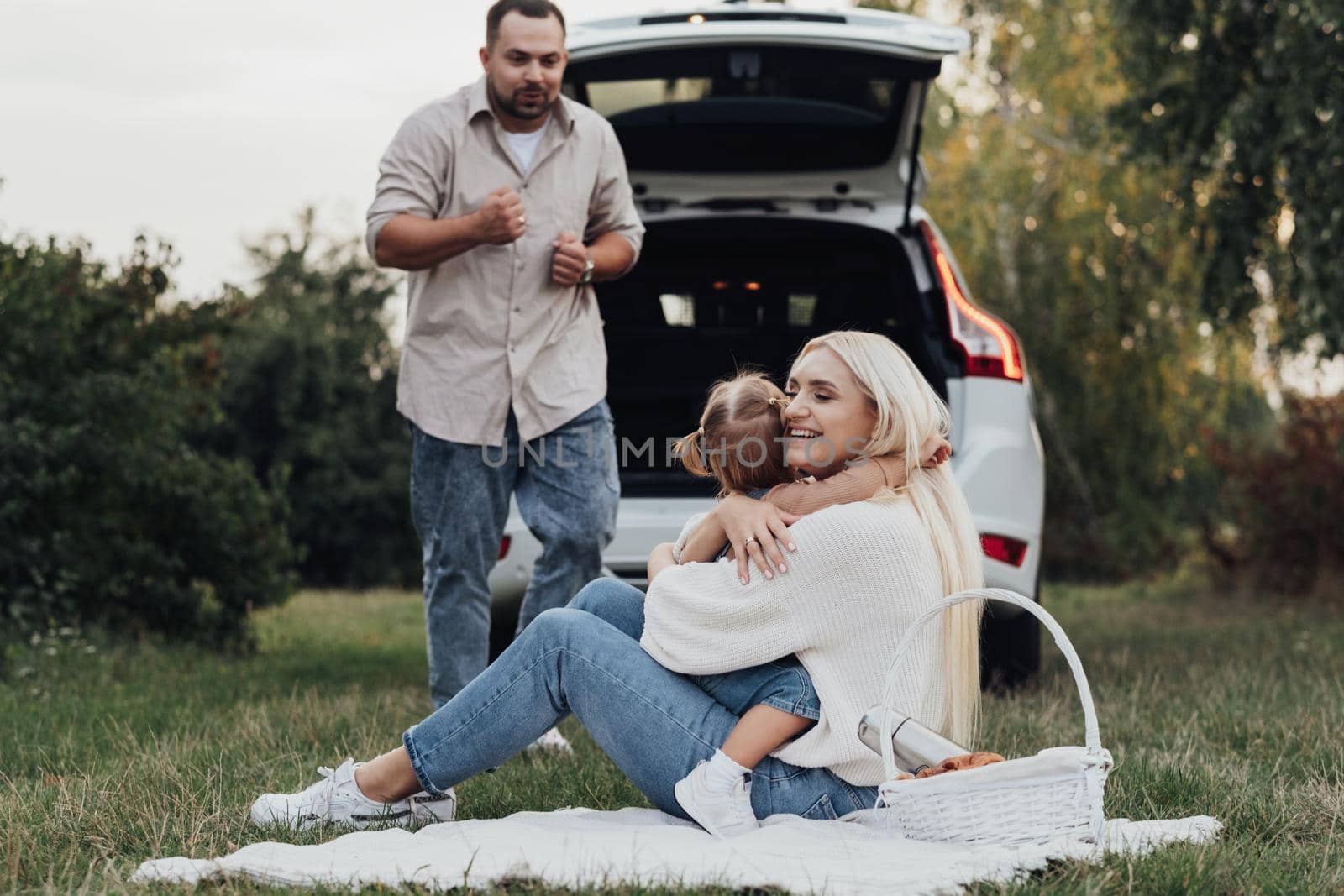 Young Mother Hugging with Her Little Daughter While Father Dancing Near Them, Family Enjoying Picnic Outdoors