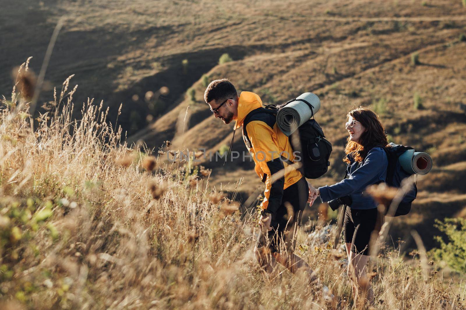 Travel Couple Man and Woman with Backpacks and Camping Mat Holding by Hands Moving on the Hill During Sunset by Romvy