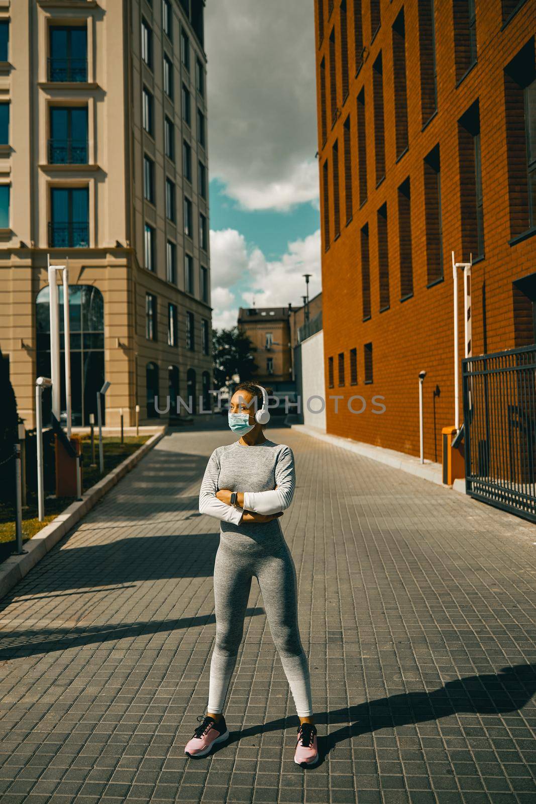 Motivated sportswoman in sportswear and mask listening to music via headphones, standing in the city