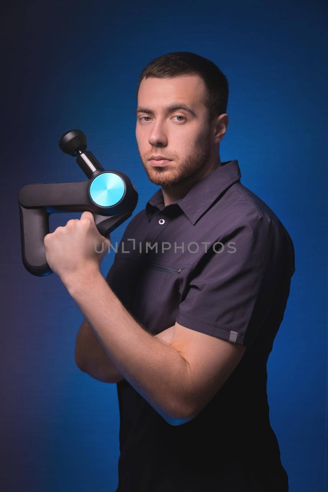 Portrait of a confident professional masseur with a percussion massager in his hands. Low key, blue background. Shock wave massage by yanik88
