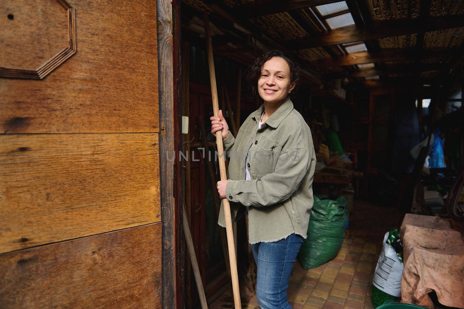 Happy female gardener farmer with gardening tools standing in her wooden rural workshop, enjoying gardening on a beautiful early spring day by artgf