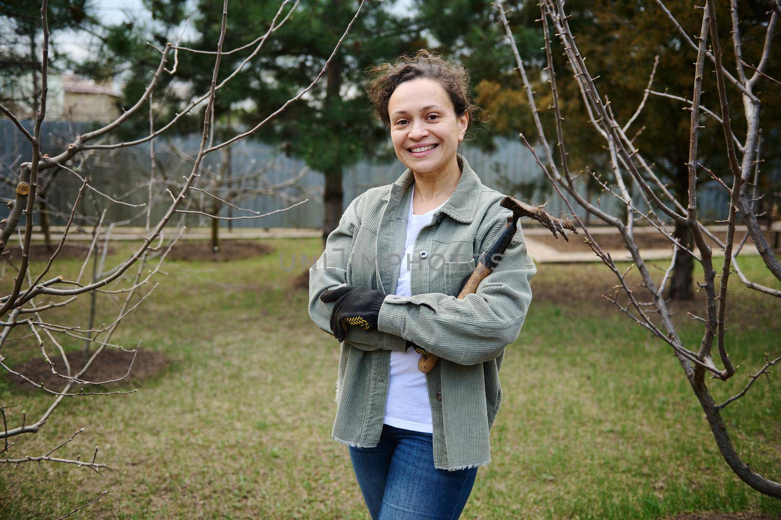 Happy female gardener holding a soiled rake in her hands and smiles looking at camera, standing in her countryside botany garden