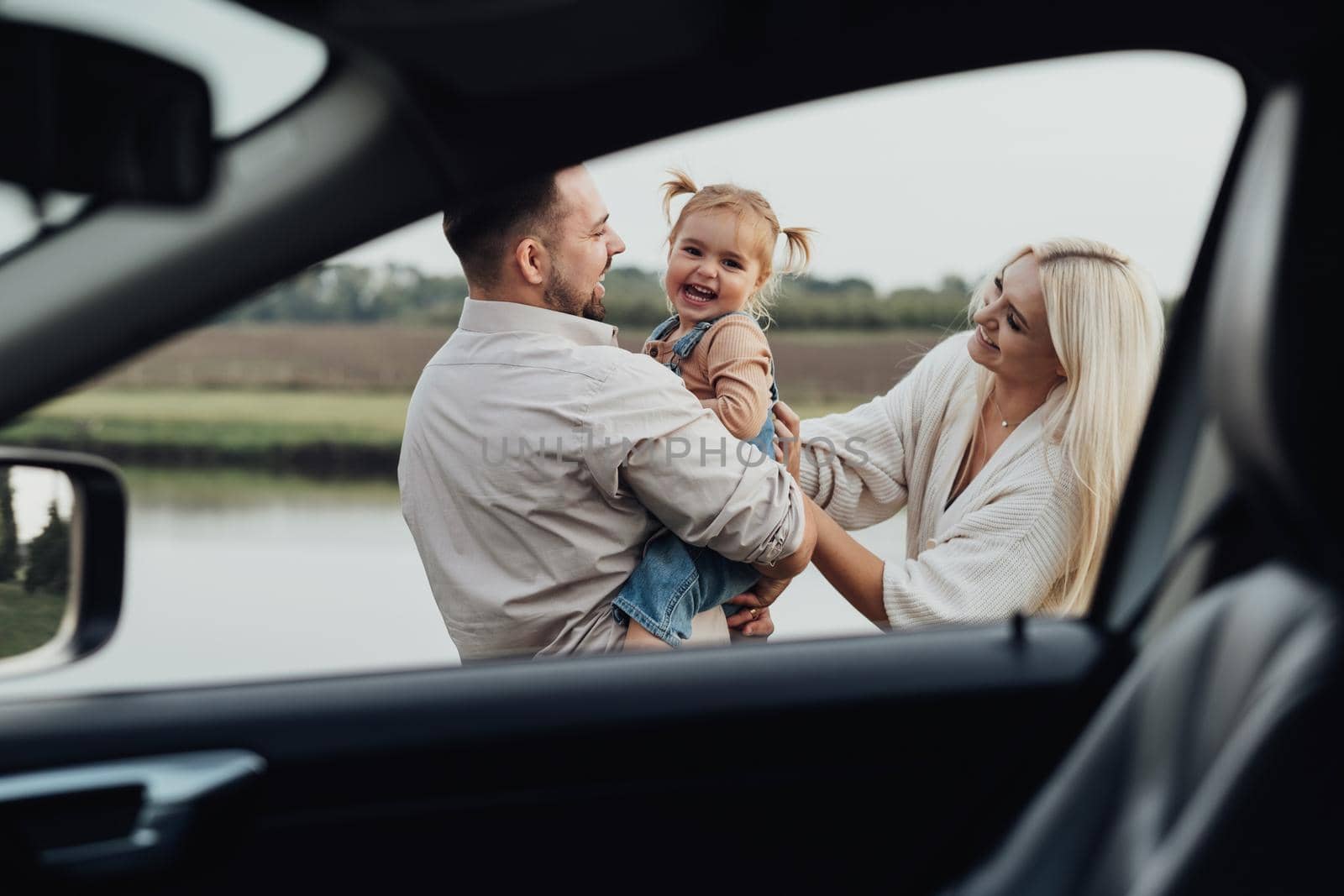 View Through Window Inside of Car, Happy Young Family, Mom and Dad with Their Little Daughter Having Fun Together and Enjoying Weekend Outside the City by Romvy