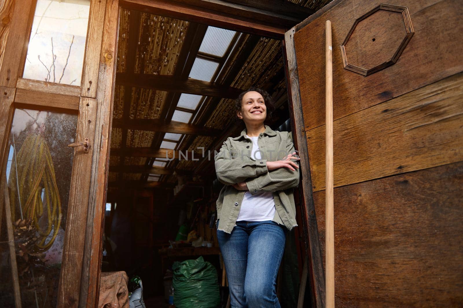 View from the bottom to a serene confident female gardener standing with crossed arms in her wooden countryside workshop with gardening tools and smiling looking at camera by artgf