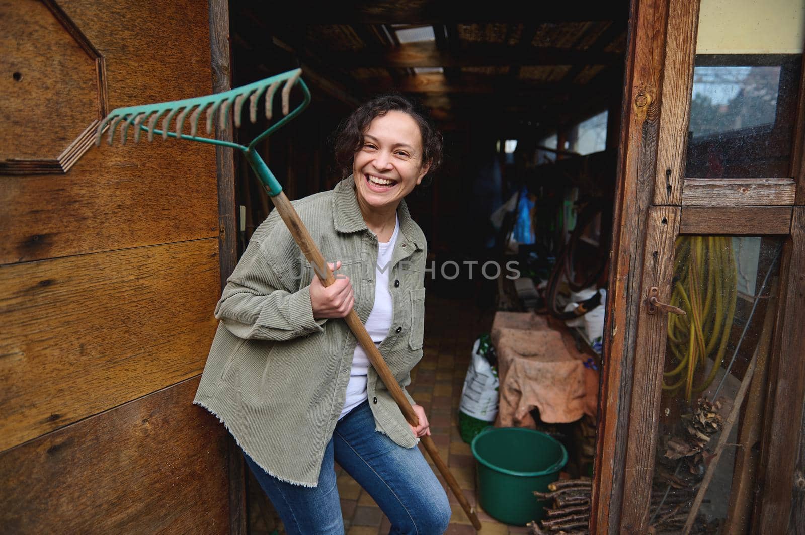 Happy pleasant mature female gardener holding a rake and smiling with a cheerful toothy smile while standing in her wooden rural workshop with gardening tools enjoying home gardening on a spring day