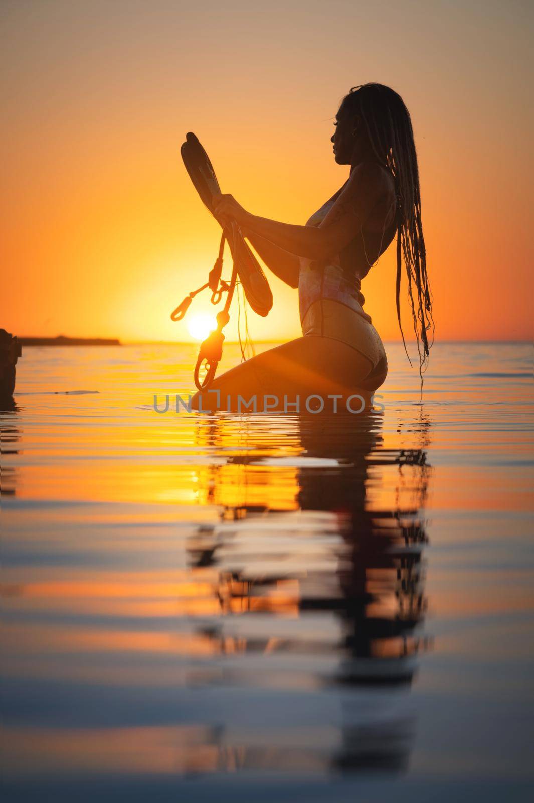 Silhouette frame. A young slim sexy female kitesurfer with a plank and a kiteboard stands in the water in the shallow water at sunset. Water sports. Stylized frame.