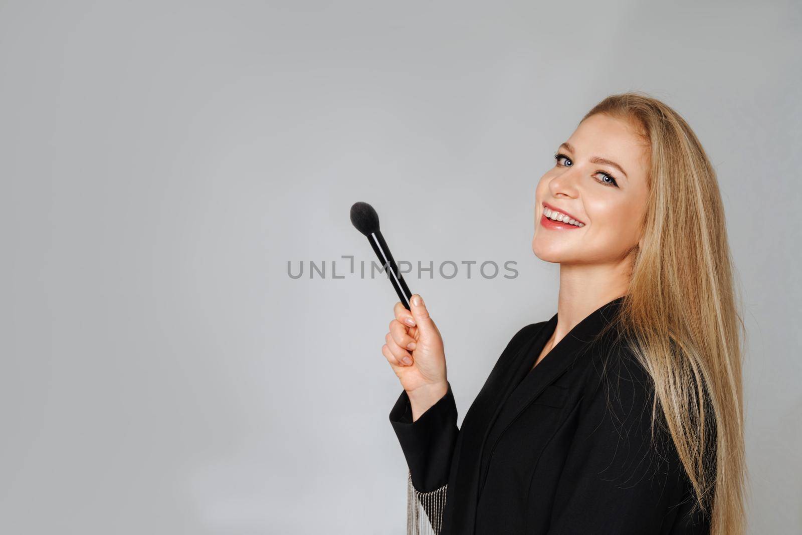 Beautiful middle aged woman makeup waving one makeup brush, winking at the camera and smiling. Blond hair and a black jacket on a light background