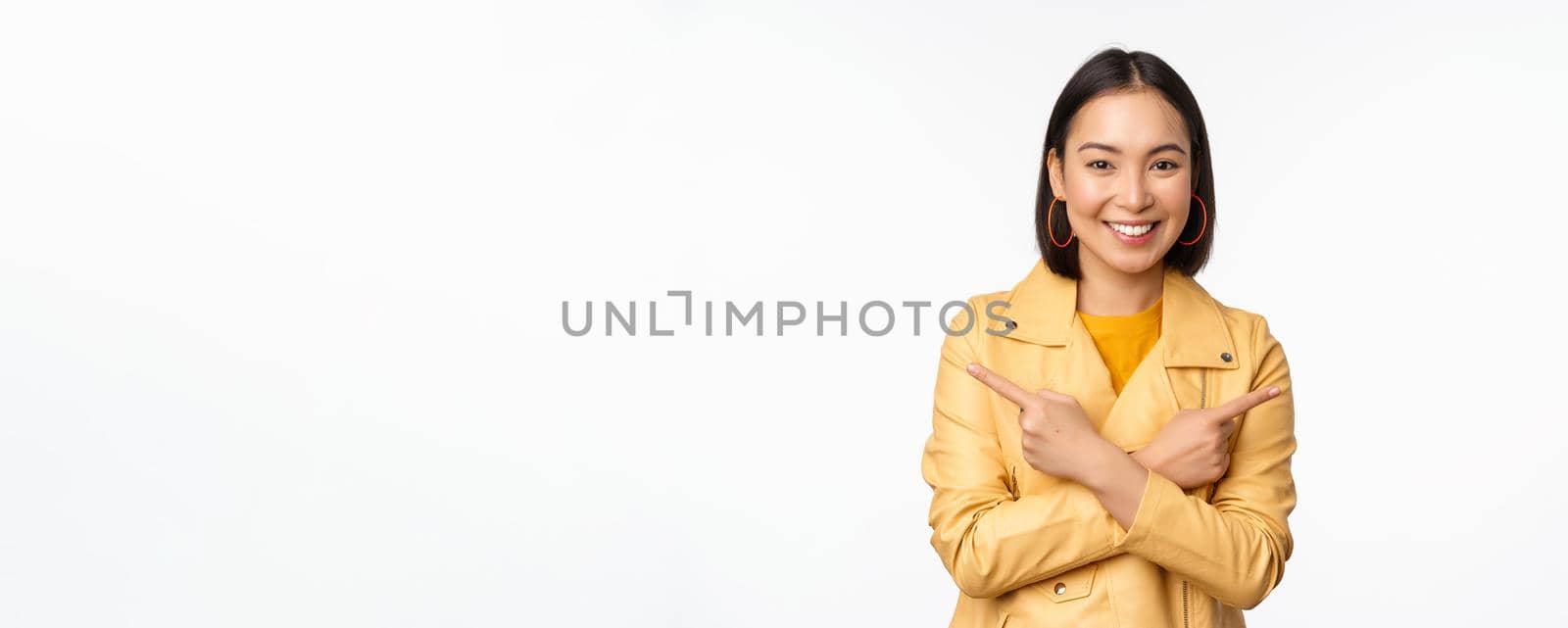 Image of indecisive asian girl, pointing fingers sideways, pointing left and right, choosing variant, deciding, standing over white background. Copy space