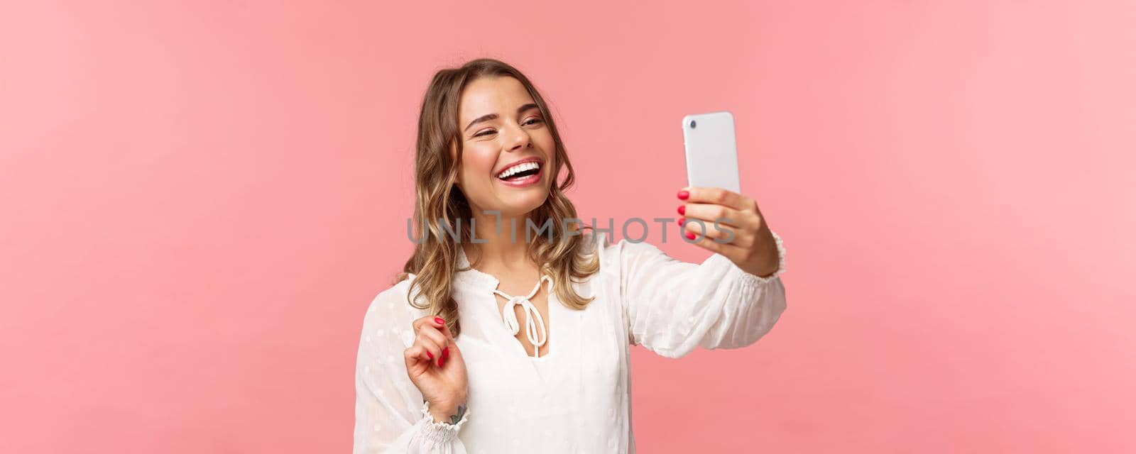 Close-up portrait of cheerful upbeat smiling blond girl, wearing white dress, laughing as record video, calling friend on mobile application, taking photo, selfie on smartphone, pink background.