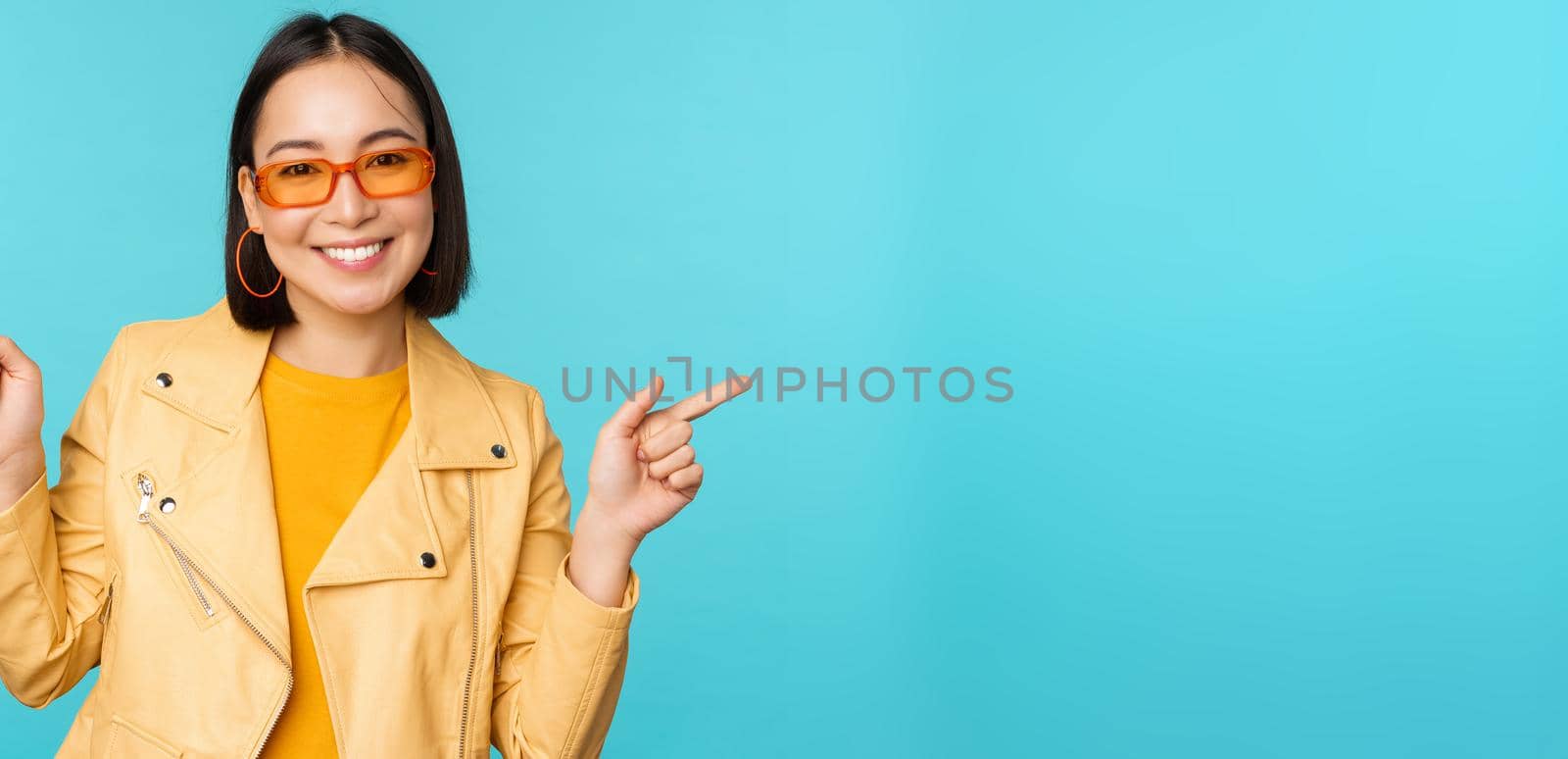 Happy stylish chinese girl in sunglasses, points fingers sideways, invites to check out, see store discount, shows left and right, stands over blue background.