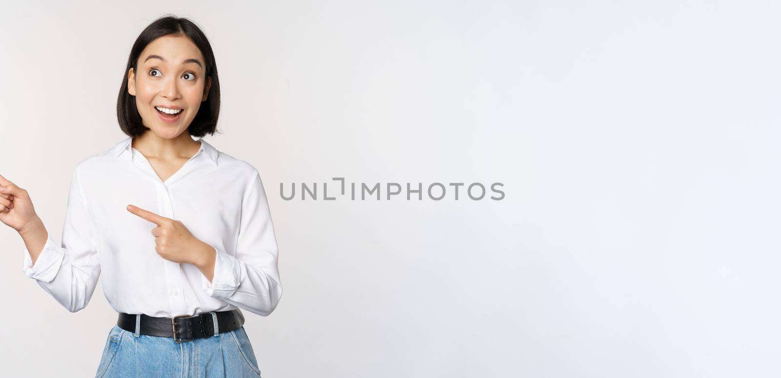 Enthusiastic korean girl pointing fingers left, female student pointing and looking left with happy smile, showing company logo or banner, white background by Benzoix