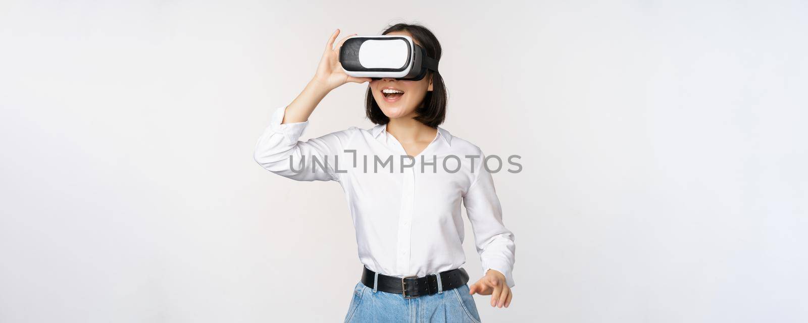 Excited young woman enter virtual reality in her glasses. Asian girl using vr headset, standing over white background by Benzoix