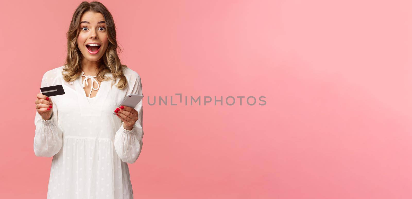 Portrait of excited and amused, happy smiling caucasian woman making order internet, shopping online with mobile phone, enter credit card, billing info, look camera amazed, pink background by Benzoix