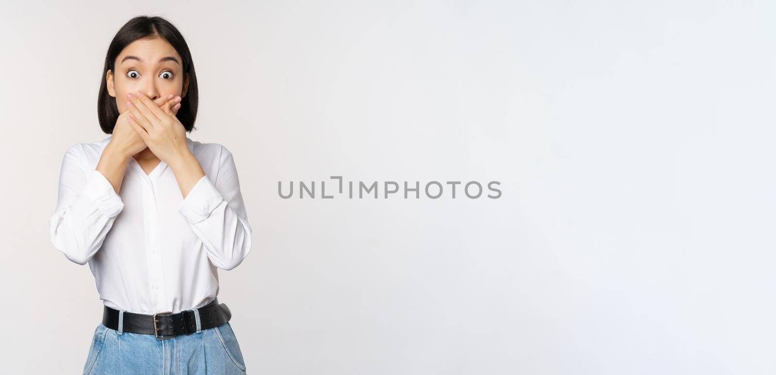 Shocked young asian girl close mouth, cover lips with hands and looking amazed, standing over white background. Taboo gesture