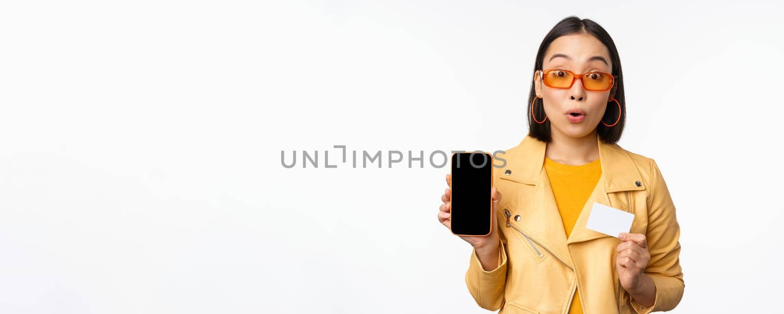 Online shopping and people concept. Stylish asian woman showing mobile phone screen and credit card, smartphone application, standing over white background by Benzoix