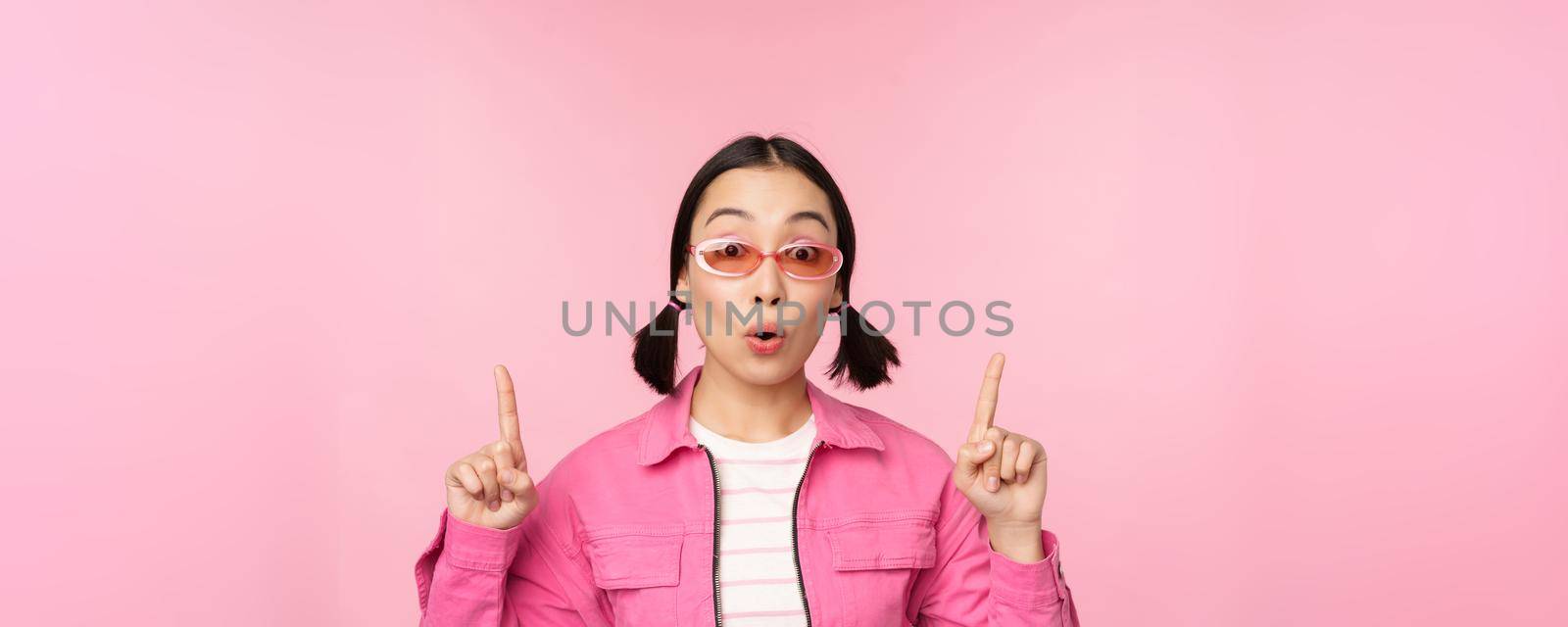 Close up portrait of asian woman gasping, looking surprised, pointing finger at banner, advertisement, standing over pink background.