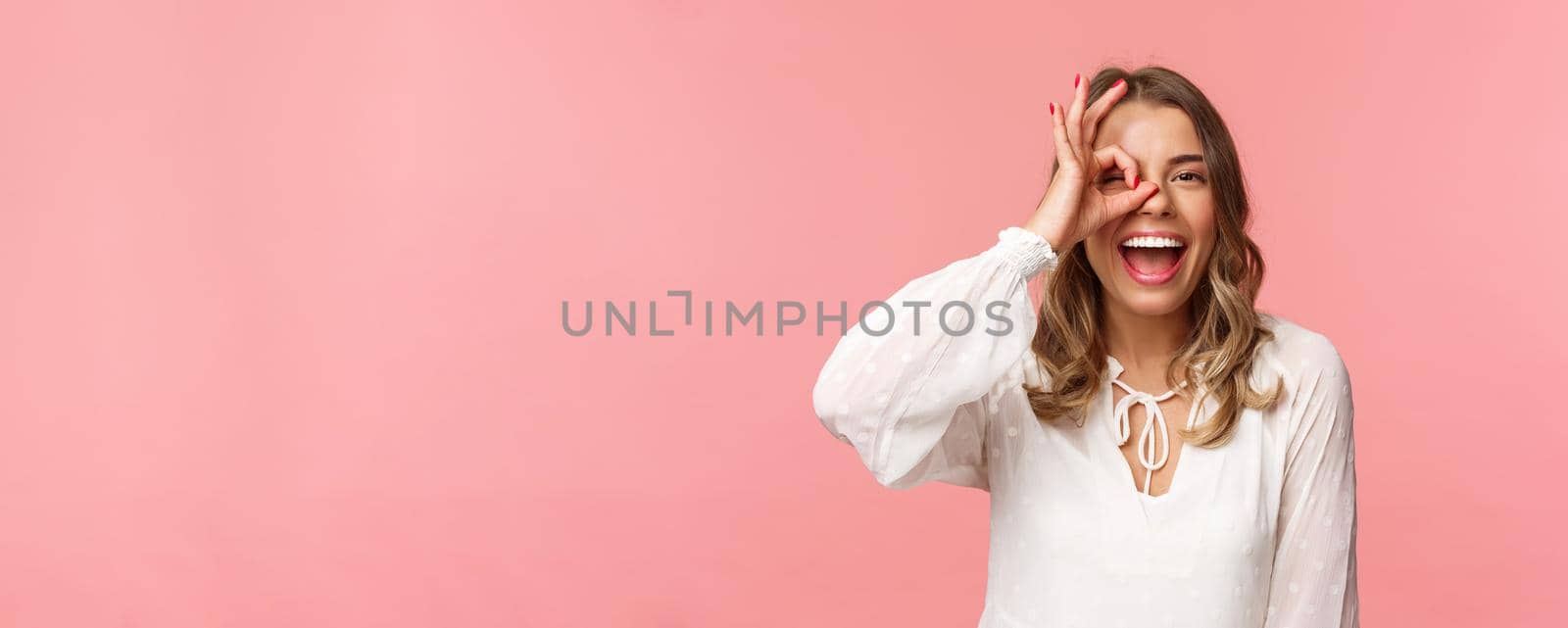 Close-up portrait of perfect good-looking blond girl in white dress, looking through okay sign and smiling, guarantee excellent quality, approve or like something, standing pink background.
