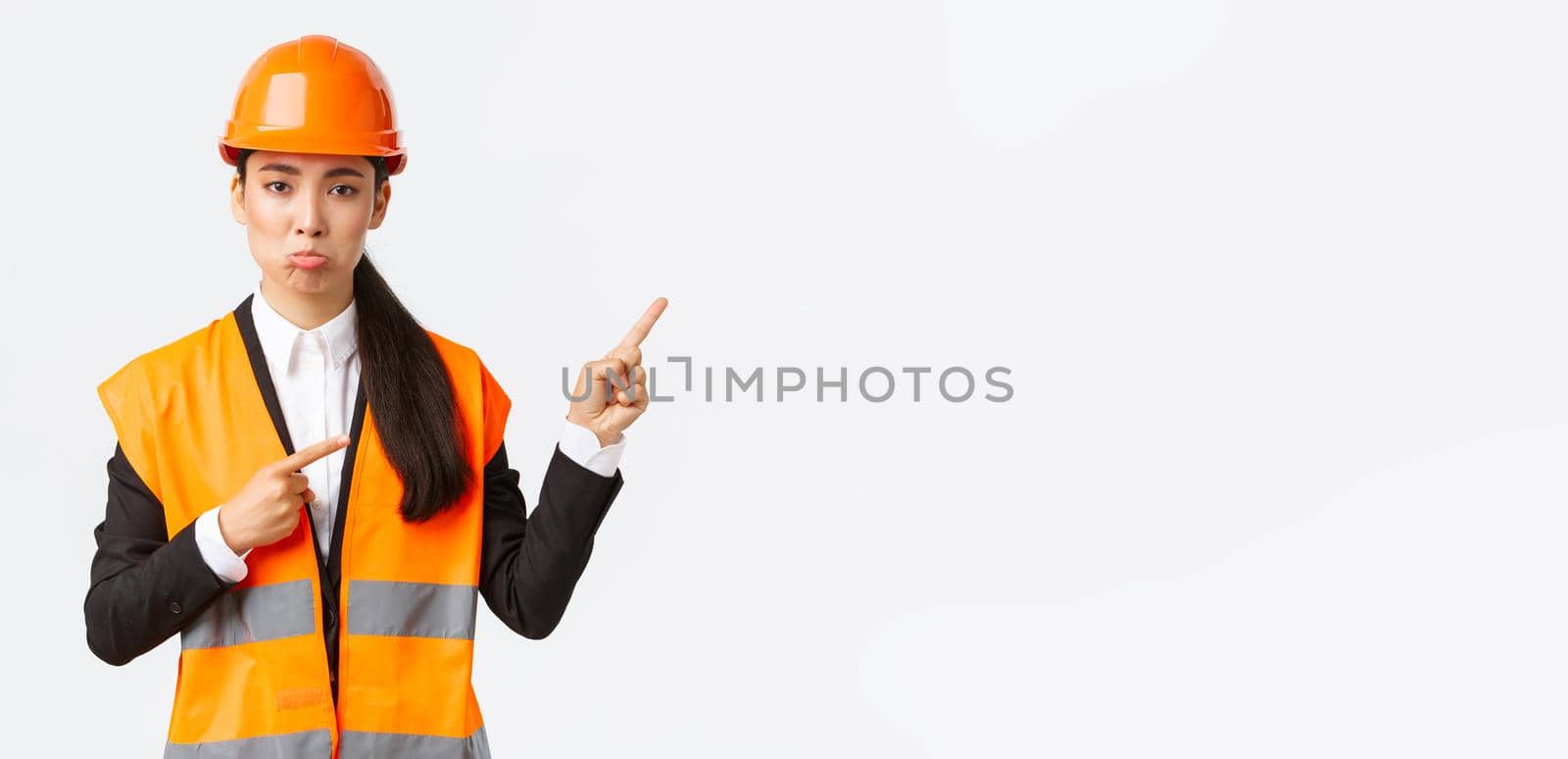 Building, construction and industrial concept. Upset and disappointed female asian manager, engineer in safety helmet pouting, complaining something bad, pointing fingers upper right corner.