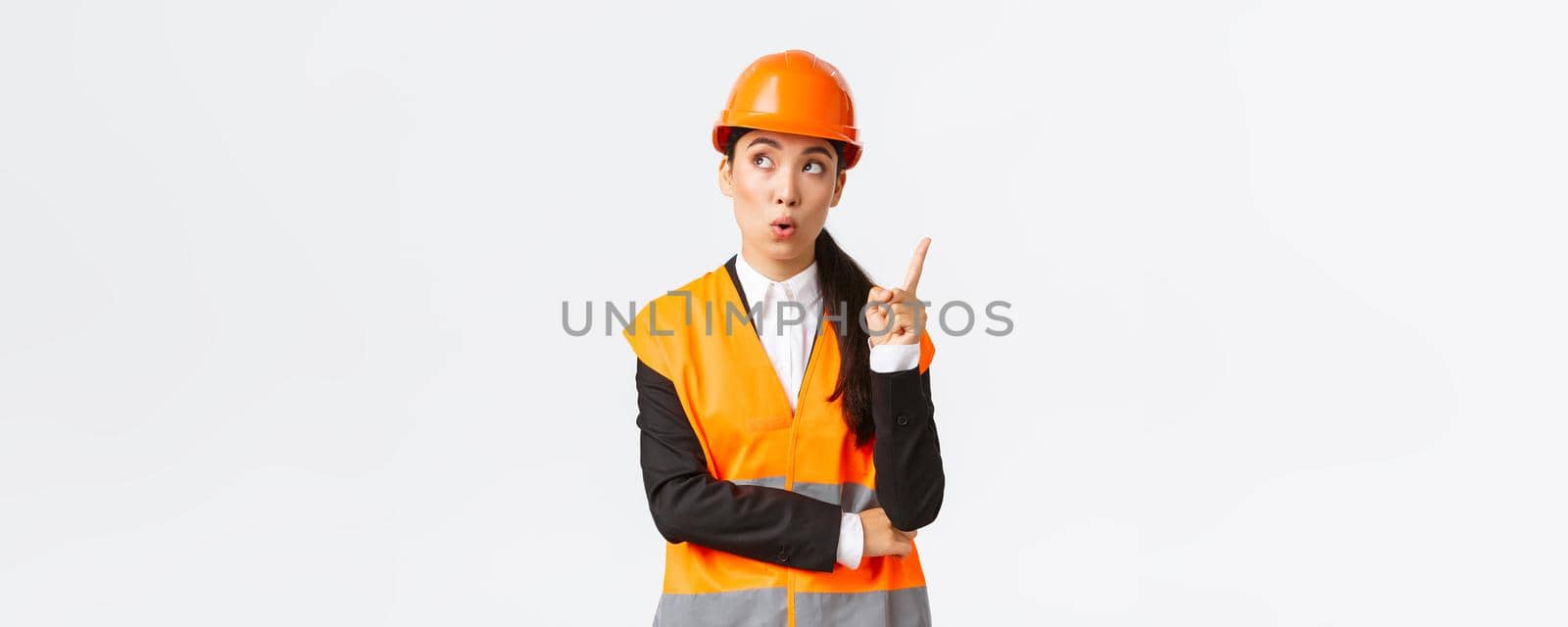 Thoughtful creative female asian engineer, architect in safety helmet and reflective jacket, raising index finger, eureka gesture, have interesting idea, suggest plan, have solution, white background.