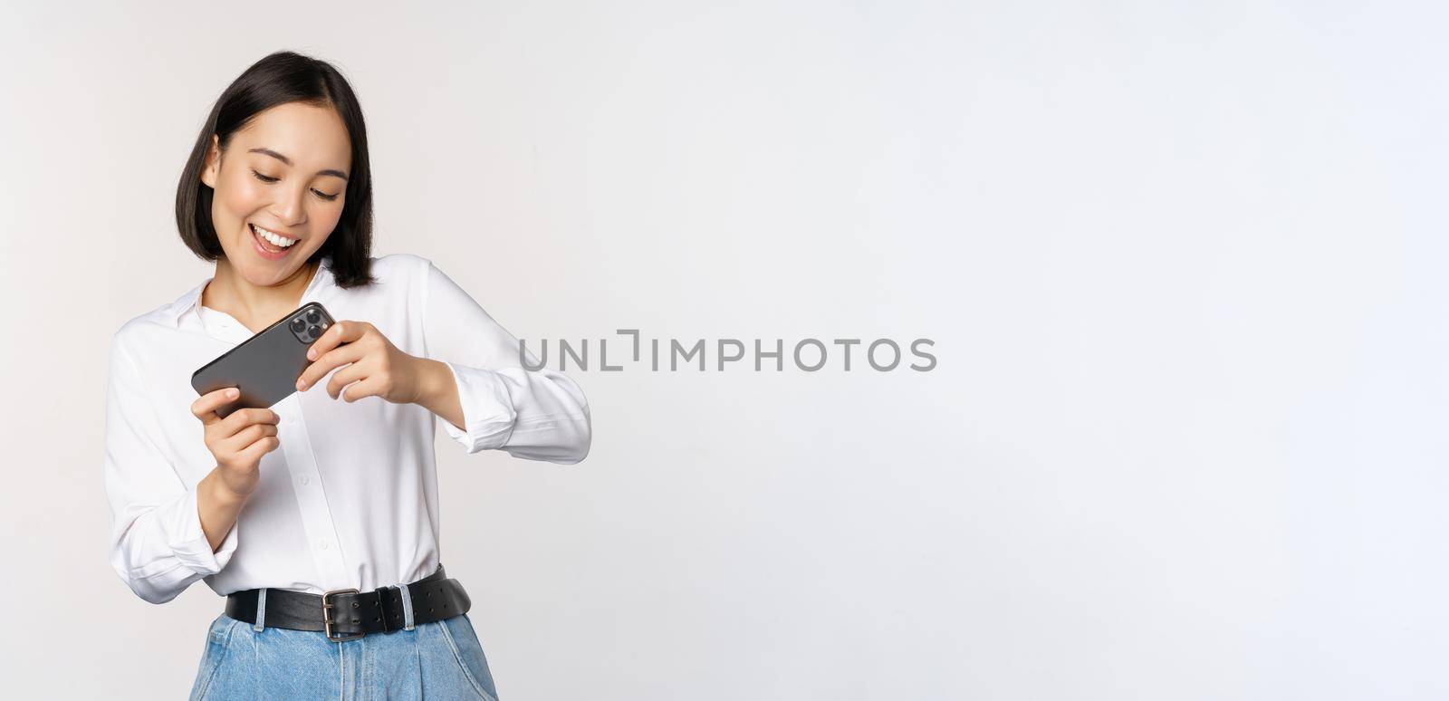 Young korean woman, asian girl playing mobile video game on smartphone, looking at horizontal phone screen, standing over white background by Benzoix