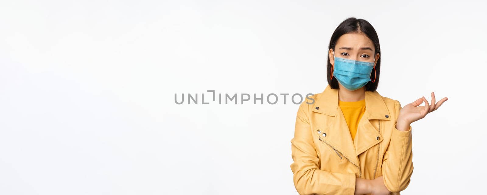 Confused asian woman in medical face mask, looking clueless, puzzled, wearing medical face mask, standing over white studio background.