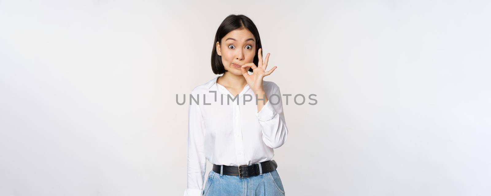 Mouth shut. Young asian woman seal lips, showing taboo dont speak, keep quiet sign, hiding secret, standing over white background.