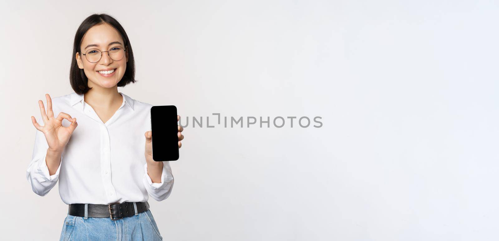 Image of asian businesswoman, showing smartphone screen, app interface and ok sign, recommending application on mobile phone, standing over white background.