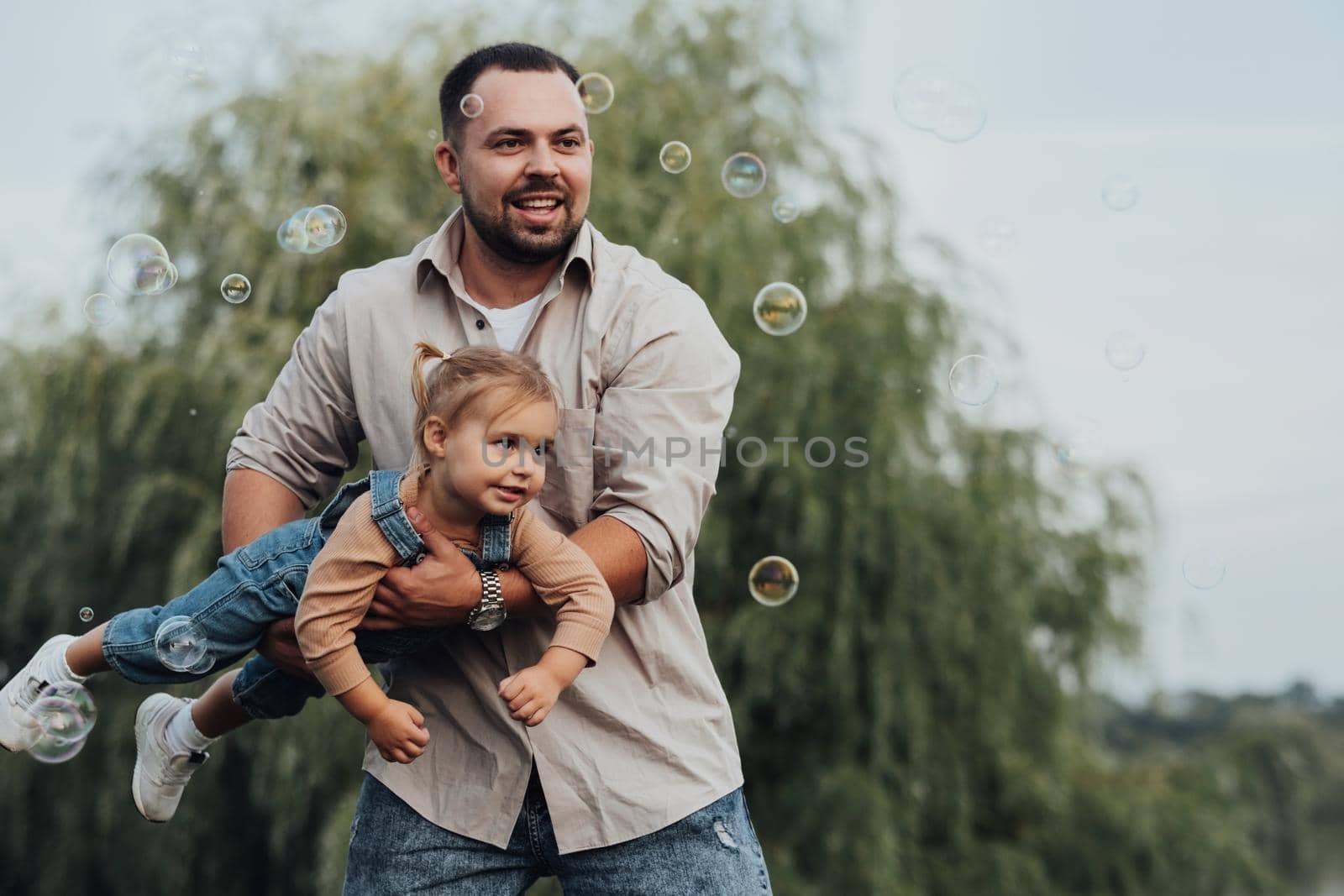 Happy Young Father Holding His Daughter in Arms and Imitating Flying and Playing with Bubbles Outdoor by Romvy
