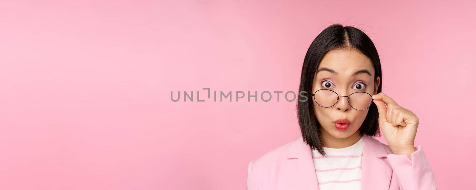 Close up portrait of korean office lady takes off glasses and looking impressed at camera, surprised face expression, pink background.