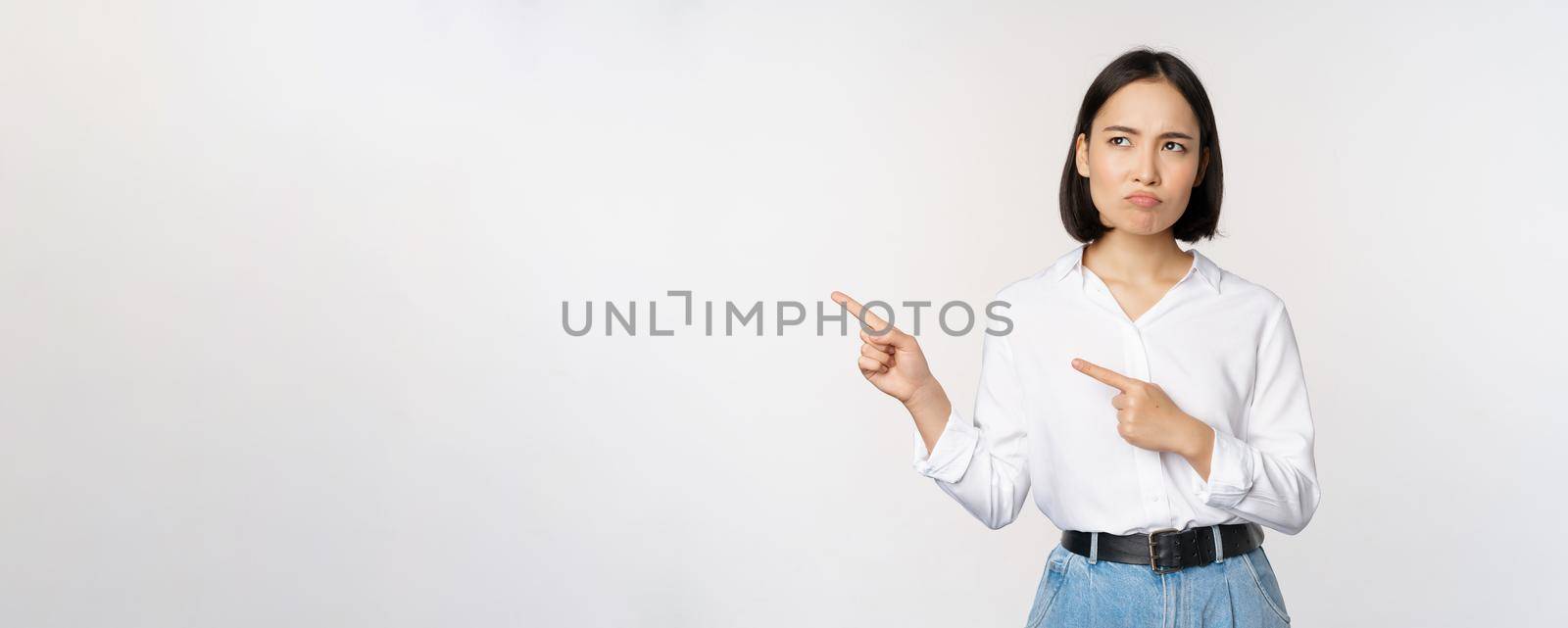 Disappointed, sad young asian woman pointing and looking left with upset, sulking face expression, standing over white background.