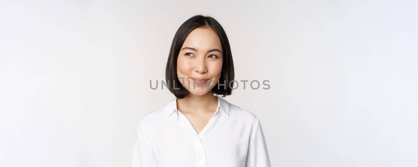 Image of smiling asian woman planning, thinking of smth, daydreaming, standing over white background with smug face. Copy space