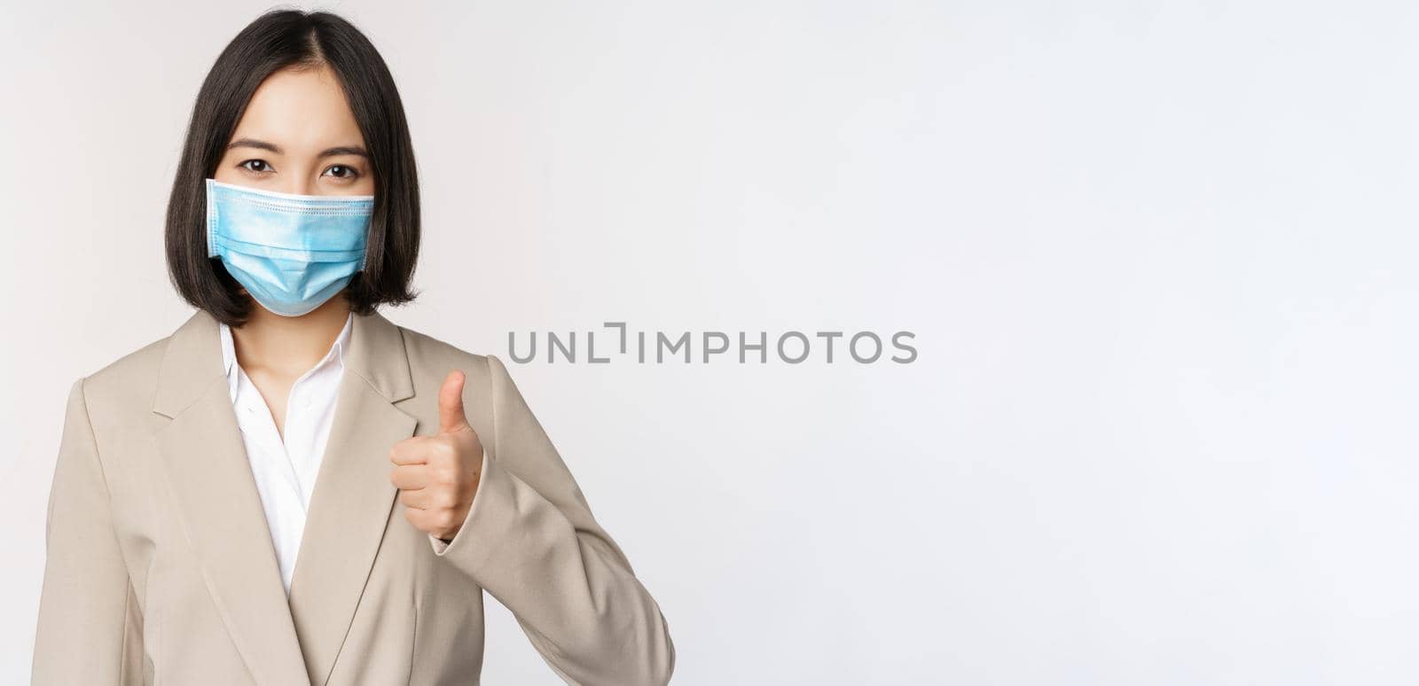 Coronavirus and workplace concept. Portrait of businesswoman in medical face mask showing thumbs up, wearing business suit, standing over white background.