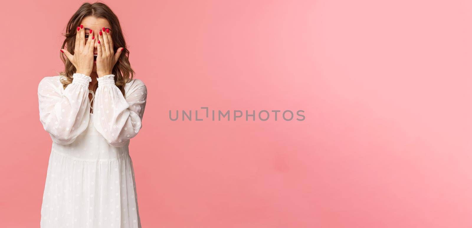 Portrait of silly cute girl with blond short hair, wear white dress, hiding face behind hands, promise to wait for signal but peeking through fingers, cant resist temptation, stand pink background.