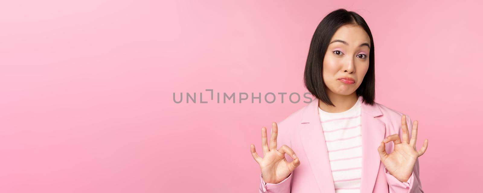 Not bad. Impressed businesswoman, asian office lady showing okay sign and nod in approval, standing over pink background. Copy space