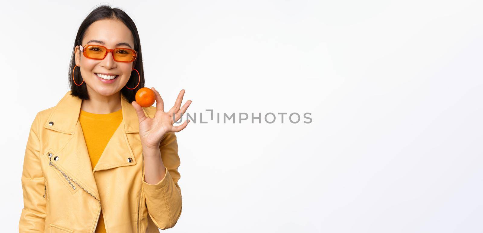 Beautiful asian girl in sunglasses showing tangerine and smiling, looking happy, posing in yellow against studio background.