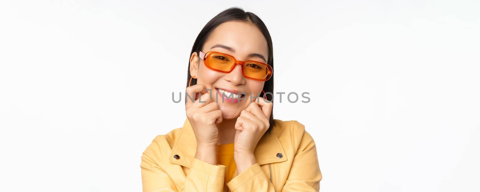 Close up portrait of trendy asian woman in sunglasses, touching her face, looking romantic, smiling at camera, standing over white background by Benzoix