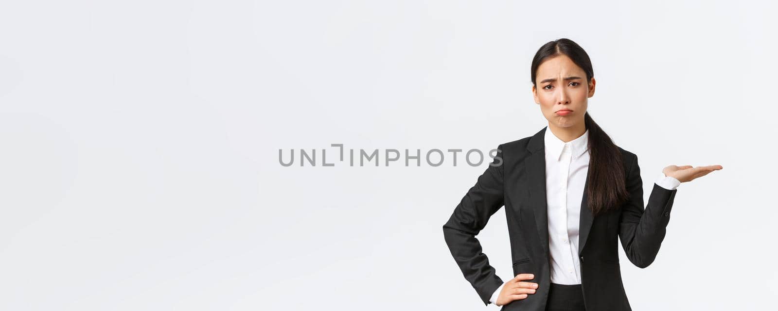 Disappointed and gloomy female sales manager, real estate agent in black suit having bad day, pouting and look upset while showing something, holding hand to the right, white background.