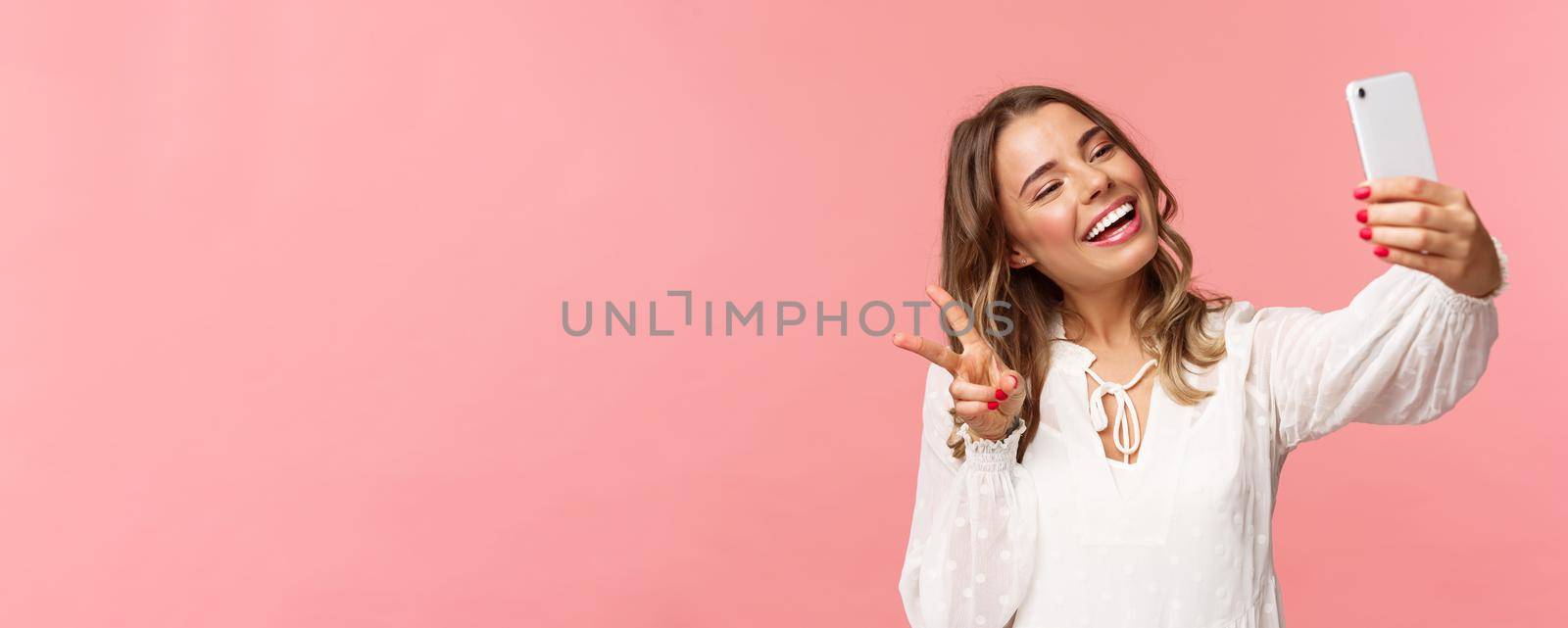 Close-up portrait of cheerful lovely, feminine blond girl in white dress, taking selfie on mobile phone, make kawaii peace sign while take photo, capturing spring moment, pink background.